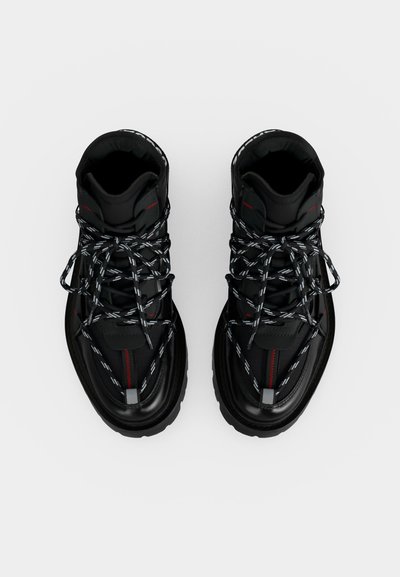 Paire de bottes montantes noires avec semelles épaisses et lacets noirs et blancs à motifs vues de dessus sur fond blanc.