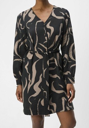 Femme portant une robe noire boutonnée avec des motifs ondulés abstraits beige et une ceinture en tissu assortie nouée à la taille.