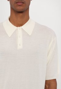 Polo crème avec un col tricoté, doté d'une patte de boutonnage à trois boutons et de manches courtes, fabriqué dans un tissu léger et texturé.