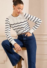 Maglione a righe in crema con dettagli blu navy e oro, con bordi scallop. Abbinato a jeans svasati blu scuro e uno sgabello di legno.