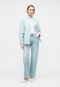 Frau trägt hellblaue Jeansbluse und Jeans, weißes Tanktop, weiße spitze High Heels, steht mit einer Hand in der Tasche vor weißem Hintergrund.