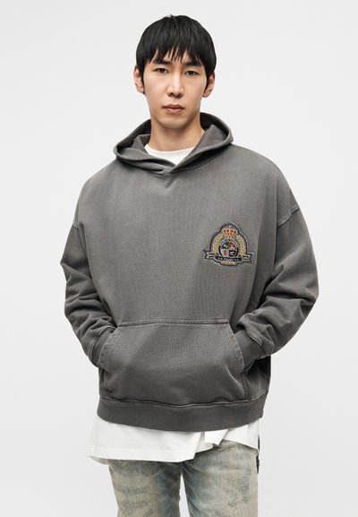 Hall halliga kapuutsiga dressipluus suure tikitud embleemiga rinnal. Omab esist taskut ja lõdvestatud lõiget koos soonikuga mansettidega.