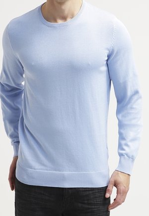 Homme portant un pull à col rond à manches longues bleu clair et un pantalon noir, se tenant debout avec les bras relâchés sur les côtés.
