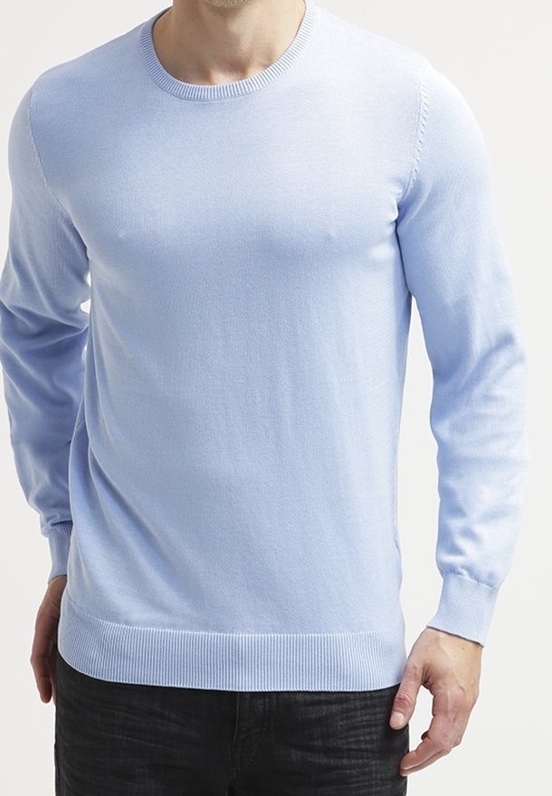 Uomo che indossa un maglione a maniche lunghe di colore azzurro chiaro e pantaloni neri, in piedi con le braccia rilassate ai lati.