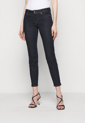 Vaqueros ajustados azul oscuro combinados con sandalias negras de tacón alto con tiras, en una persona con fondo liso.