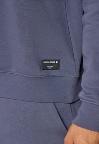 Σκούρο μπλε φούτερ με ριμπ μανσέτες και στρίφωμα. Διαθέτει μια μαύρη ετικέτα μάρκας με λευκό κείμενο στη χαμηλότερη πλευρά. Μαλακό, υφασμάτινο ύφασμα.