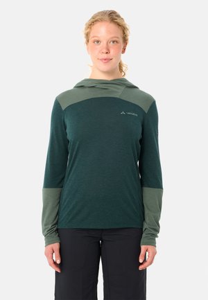 Frau trägt einen zweifarbigen grünen langärmligen Kapuzenpullover und schwarze Hose, steht frontal vor einem schlichten Hintergrund.