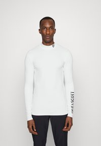Long-sleeve white performance shirt with a high collar, featuring a logo on the neck and branding along the left sleeve. Smooth texture.