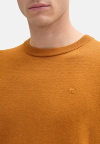 Oranger Strickpullover mit klassischem Rundhalsausschnitt. Verfügt über ein kleines Logo-Detail auf der Brust. Glatte Textur, mittelschwere Stoffqualität.