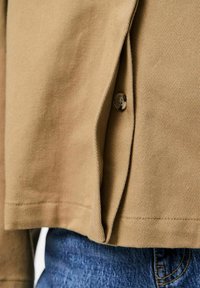 Rjav, teksturiran srajc s gumbom na strani, v kombinaciji z modrimi jeans hlačami. Tkanina prikazuje diagonalen vzorec tkanja.