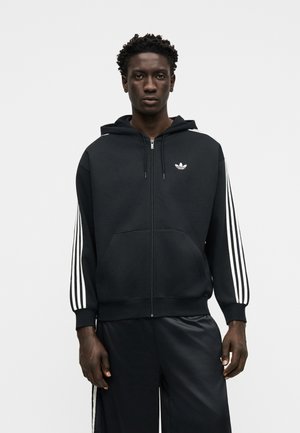 Mann steht und trägt einen schwarzen Adidas Kapuzenpullover mit Reißverschluss und weißen Streifen an den Ärmeln sowie passende schwarze Hosen mit Seitenstreifen.