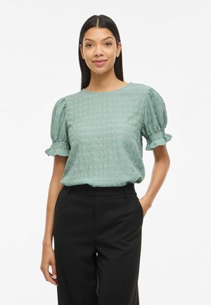 Kobieta o długich ciemnych włosach, ubrana w teksturowaną bluzkę z bufiastymi rękawami w kolorze szałwii oraz czarne dopasowane spodnie, stojąca z jedną ręką w kieszeni.