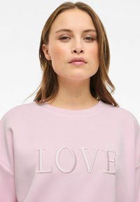 Sweatshirt rose clair avec un col rond et des épaules tombantes ; arbore un "LOVE" brodé dans un rose légèrement plus foncé. Texture douce.