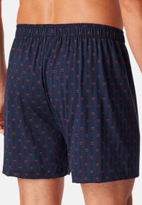 Marineblauwe boxershorts met een elastische tailleband, versierd met een herhalend geometrisch patroon van rode en zwarte accenten. Zachte stoftextuur.