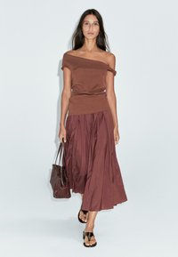 Top marrone scollato con texture arricciata abbinato a una gonna marrone fluida e plissettata, e una borsa coordinata. Il modello indossa sandali neri.