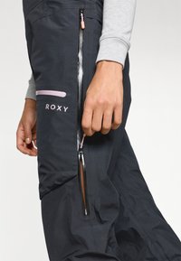 Calças de neve pretas com detalhes em ziper rosa contrastantes e marca. Apresenta um bolso com fecho e um design elegante. O material parece leve.