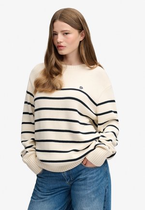 Femme aux longs cheveux bruns portant un pull crème à rayures noires et un jean bleu, debout les mains dans les poches devant un fond uni.