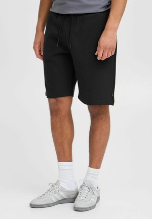 Pantaloni shorts neri texture con vita elastica e coulisse, tasche laterali, abbinati a sneaker grigie e calze bianche.