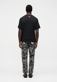 Μαύρο πουκάμισο με κοντά μανίκια και μικρό κόκκινο λογότυπο στην πλάτη, συνδυασμένο με γκρι παντελόνια με σχέδιο. Εικόνα από πίσω.