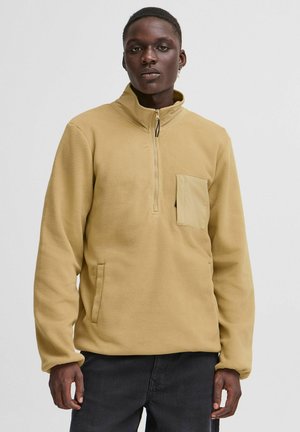 Light brown fleece pullover with a high collar, zippered front, and a chest pocket. Features an overall soft texture and loose fit.