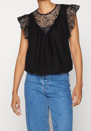 Kvinde iført en sort bluse med blonder og flagrende ærmer samt halsudskæring, kombineret med højtaljede blå jeans, vist fra hals til hofter.