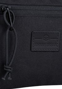 Pochette en tissu noir avec surface texturée, dotée d'une fermeture éclair, d'une tirette noire et d'un patch en relief portant le logo "Johnny Urban".