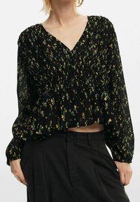 Blouse florale noire en tissu léger. Elle présente un col en V, une taille froncée et des manches longues avec des poignets élastiques. Motif floral jaune et vert.