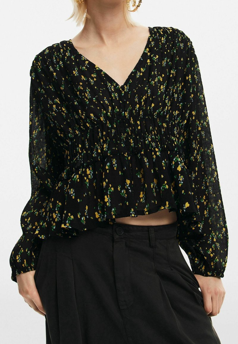 Blouse florale noire en tissu léger. Elle présente un col en V, une taille froncée et des manches longues avec des poignets élastiques. Motif floral jaune et vert.