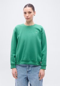 Sweat-shirt vert avec col rond, poignets et ourlet côtelés. Présente un petit logo sur la poitrine. Associé à un jean bleu clair.