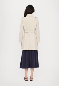 Trench coat bej cu talie ajustată prin curea, mâneci lungi și fante la spate, asortat cu o fustă bleumarin până la genunchi și sandale cu curele.
