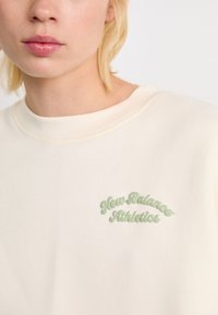 Blonde persoon die een crèmekleurige sweatshirt draagt met lichtgroene tekst "New Balance Athletics" op de borst.