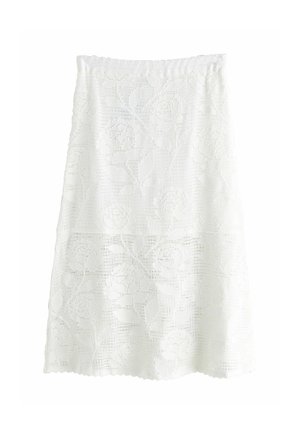 Młoda kobieta w białym topie bez rękawów i długiej koronkowej spódnicy trzyma plecioną torebkę, stojąc na drewnianej podłodze blisko kamiennej ściany i okna.