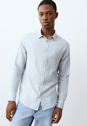 Giovane uomo con indosso una camicia grigia chiaro con bottoni e maniche lunghe e jeans blu, in piedi contro uno sfondo bianco uniforme.