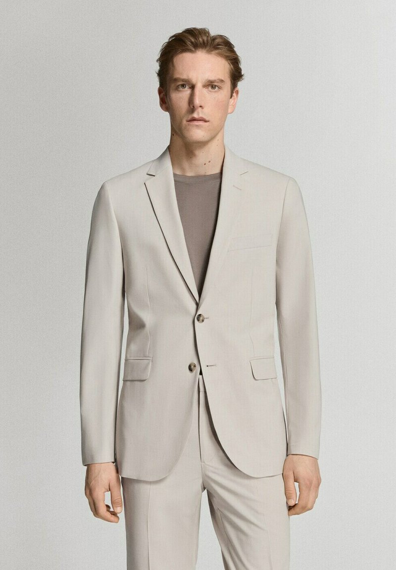 Hombre con cabello corto y castaño, vestido con chaqueta y pantalones de traje beige claro sobre una camisa topo, de pie frente a un fondo liso y claro.