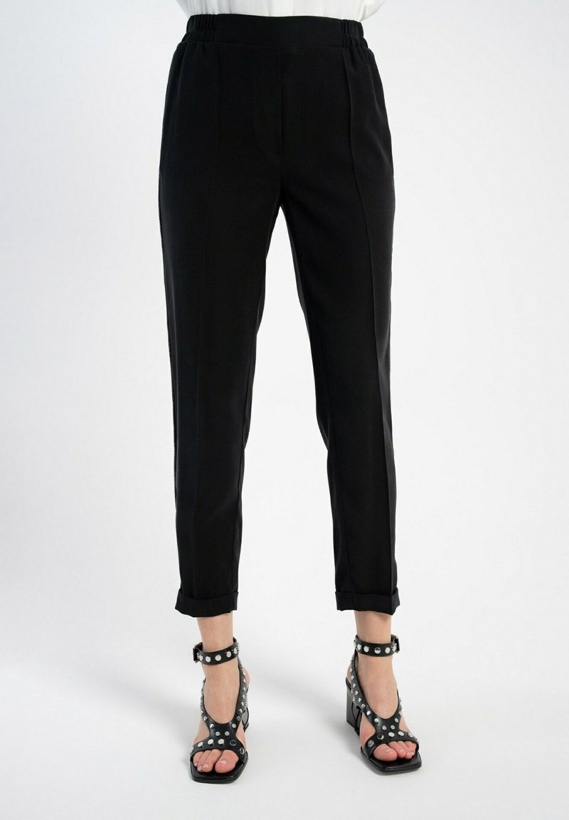 Czarne spodnie o zwężanym kroju z elastycznym pasem, z dwiema fałdami z przodu i podwiniętymi mankietami, zestawione z czarnymi sandałami na obcasie z odsłoniętymi palcami, ozdobionymi srebrnymi ćwiekami.
