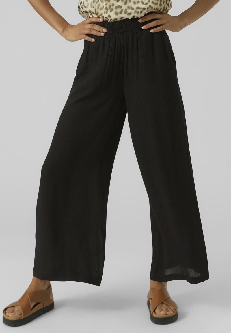 Femme portant un pantalon noir à jambes larges, un haut à imprimé léopard et des sandales compensées marron, debout les mains sur les hanches.