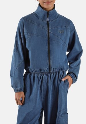 Frau trägt eine blaue Reebok-Jacke mit Reißverschluss und elastischem Bund sowie passende Hose, Hand in der Tasche, vor einfachem Hintergrund.