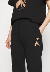 Camiseta de algodón negra con un estampado gráfico en bronce, combinada con pantalones negros que cuentan con bolsillos laterales y un detalle de logo en bronce a juego.