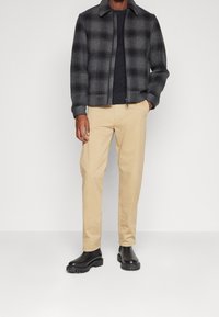 Man som bär en svart och grå rutmönstrad jacka, mörk tröja, beige byxor och svarta kraftiga stövlar mot en enfärgad bakgrund.