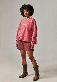 Rosa hettegenser med grafisk stjernedesign, paired med røde rutete shorts. Modellen har på seg brune ankelstøvler. Bakgrunnen er nøytral grå.