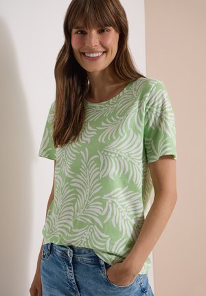 Femme souriante portant une chemise à manches courtes vert clair avec des motifs de feuilles blanches et un jean bleu, debout avec une main dans sa poche.