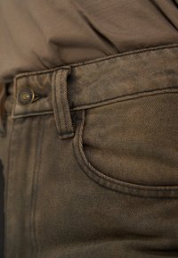 Κοντινή λήψη της ζώνης μέσης καφέ τζιν με θηλιά ζώνης, κουμπί και μπροστινή τσέπη, σε συνδυασμό με ένα ταιριαστό καφέ πουκάμισο που είναι στολισμένο μέσα στο παντελόνι.