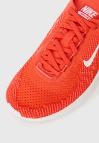 Piros hálós sportcipő, texturált felülettel, lapos fűzőkkel és fehér Nike pipa logóval. Kerek orrú és párnázott talppal rendelkezik.