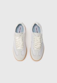 White sneakers with a textured suede upper, lace closure, and light blue insoles; brown rubber soles and subtle design accents.