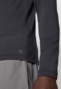 Μακρυμάνικο αθλητικό τοπ σε σκούρο γκρι ύφασμα, με εφαρμοστή γραμμή, διακριτικά ραφές και μικρό λογότυπο στο τελείωμα. Από μέσα υπάρχει ένα πιο ανοιχτό γκρι στρώμα.