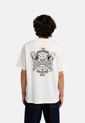 Person mit lockigem Haar trägt ein weißes T-Shirt mit schwarzem und rotem Totenkopf- und Schmetterlingsmotiv und dem Text "The Balance of Opposites" auf dem Rücken.
