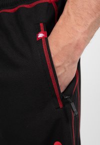 Zwarte sportieve shorts van ademend materiaal met rode accenten, een ritszak en een klein merkembleem dicht bij de zijnaad.