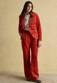 Sarkans kordurojas uzvalks ar divrindu žaketi un plašām biksēm, kombinēts ar rakstainu baltu kreklu un brūnām kurpēm.