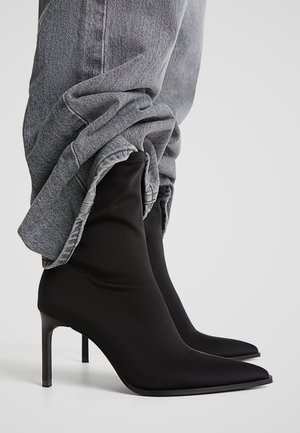 Bottes à talons aiguilles noires à bouts pointus, portées avec un jean gris retroussé, sur un fond clair uni.