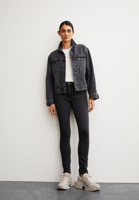 Zwarte denimjack met voorzakken, witte t-shirt, zwarte skinny jeans en lichte sneakers op een model tegen een effen achtergrond.